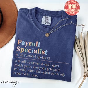 May include: Navy blue Comfort Colors t-shirt with the text "Payroll Specialist" in colorful letters. The shirt includes a definition of the job title. A gold necklace with a pendant is draped on the shirt.