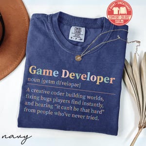 May include: Navy blue Comfort Colors t-shirt with the text "Game Developer" in colorful letters. The definition of a game developer is printed below the title. A gold necklace with a pendant is draped across the shirt.