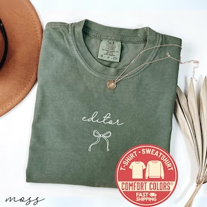 May include: A moss green Comfort Colors t-shirt with the word "editor" and a bow graphic in white. A gold necklace with a pendant rests on the shirt. A brown hat and dried leaves are in the background.
