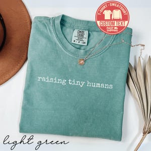 May include: A light green t-shirt with the phrase "raising tiny humans" in white lettering. The shirt has a crew neck and short sleeves. A gold-colored necklace with a pendant is draped across the shirt. A red circle with the words "T-SHIRT SWEATSHIRT" is in the upper right corner.