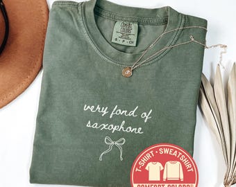 May include: A moss green Comfort Colors t-shirt with the text "very fond of saxophone" and a bow graphic. A gold necklace and a red and white sticker are also visible. The shirt is size S-P-CH.