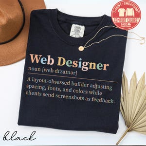 May include: Black t-shirt with the text "Web Designer" in colorful letters. The definition of a web designer is printed below the title. A gold necklace and a brown hat are also in the image.