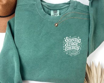 May include: Light green sweatshirt with the text "Serving Stage Energy" in white script and two theater masks. A gold necklace with a pendant is draped on the shirt. A brown hat is visible in the background.