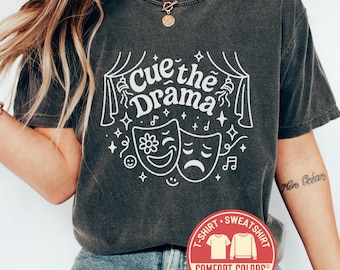 May include: Dark grey t-shirt with the text "Cue the Drama" in white, along with illustrations of theater masks, stars, and musical notes. The shirt has a crew neck and short sleeves. A red Comfort Colors logo is visible.