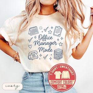 May include: Ivory-colored t-shirt with the words "Office Manager Mode" in a navy blue script, surrounded by office-themed graphics. The shirt features a Comfort Colors label and a fast shipping badge.