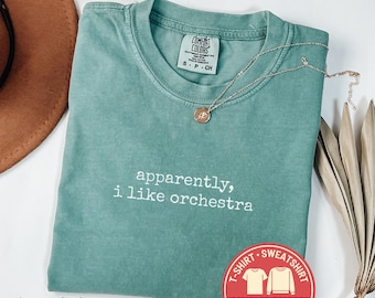 May include: A light green Comfort Colors t-shirt with the text "apparently, i like orchestra" in white. A silver necklace with a pendant rests on the shirt. A brown hat and dried leaves are in the background.