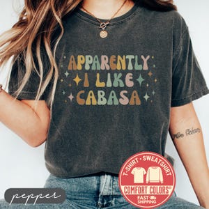May include: Dark gray Comfort Colors t-shirt with the text "Apparently I Like Cabasa" in retro-style lettering. The shirt features a round neck and short sleeves. A red and white Comfort Colors logo is visible.