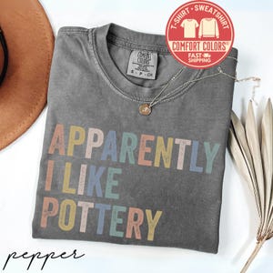 May include: A gray Comfort Colors t-shirt with the text "APPARENTLY I LIKE POTTERY" in pastel colors. The shirt has a crew neck and a small tag. A gold necklace and a hat are visible.