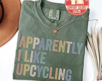 May include: An olive green t-shirt with the phrase "APPARENTLY I LIKE UPCYCLING" in colorful block letters. A gold necklace with a pendant rests on the shirt. The shirt has a Comfort Colors tag and a red sticker that says "T-Shirt Sweatshirt".