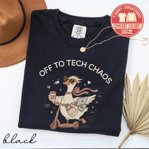 May include: A black t-shirt featuring a graphic of a goose wearing sunglasses and a scarf, riding a scooter while holding a coffee cup and a laptop. The text reads OFF TO TECH CHAOS. The shirt has a Comfort Colors label at the neck and a gold chain.