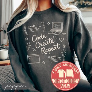 May include: Dark gray sweatshirt with the words "Code Create Repeat" in white, along with computer-related graphics. The sweatshirt has a round neck and long sleeves. A red circular logo with the words "Comfort Colors" is visible.