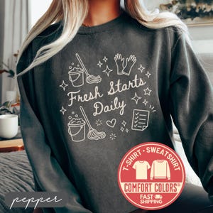 May include: Pepper-colored sweatshirt with the text "Fresh Starts Daily" and cleaning-themed graphics. The design includes a bucket, mop, gloves, and checklist. The sweatshirt has a crew neck and long sleeves.