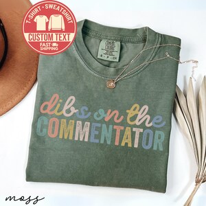 May include: A moss green t-shirt with the phrase "dibs on the COMMENTATOR" in colorful letters. A gold necklace rests on the shirt. A red circular graphic with text is in the upper left corner.