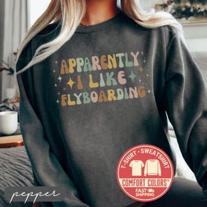 May include: Dark gray sweatshirt with the text "Apparently I Like Flyboarding" in retro-style lettering. The design includes colorful text and star accents. A red Comfort Colors logo is visible.
