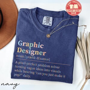 May include: Navy blue t-shirt with the text "Graphic Designer" in colorful letters. The shirt includes a definition and a quote about graphic design. A gold necklace with a pendant rests on the shirt.