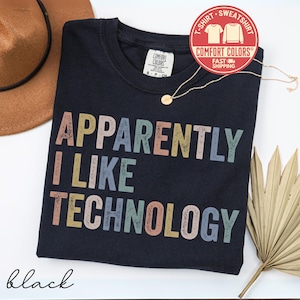 May include: Black t-shirt with the text "APPARENTLY I LIKE TECHNOLOGY" in colorful letters. The shirt has a crew neck and a Comfort Colors tag. A gold necklace and a brown hat are also visible.