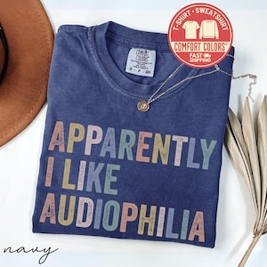 May include: Navy blue t-shirt with the text "APPARENTLY I LIKE AUDIOPHILIA" in colorful, distressed lettering. The shirt has a crew neck and short sleeves. A gold necklace with a pendant is draped over the shirt.