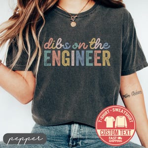May include: Dark gray t-shirt with the text "dibs on the ENGINEER" in colorful letters. A red circular graphic with the words "T-SHIRT SWEATSHIRT CUSTOM TEXT FAST SHIPPING" is visible.