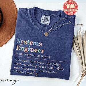 May include: Navy blue t-shirt with the words "Systems Engineer" in colorful text. The definition of a systems engineer is printed below. A gold necklace with a pendant rests on the shirt.