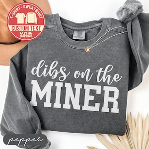 May include: A dark gray sweatshirt with the text "dibs on the MINER" in white. The sweatshirt has a crew neck and long sleeves. A necklace with a pendant is visible. The image also includes a red circle with text.