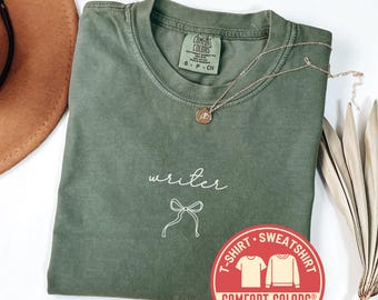 May include: Moss green Comfort Colors t-shirt with the word "writer" in cursive and a bow graphic. A gold necklace with a pendant rests on the shirt. A red sticker with "T-SHIRT SWEATSHIRT" is visible.