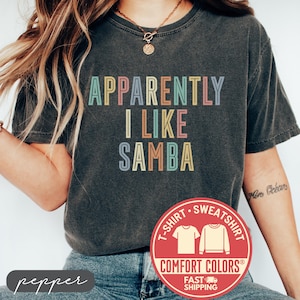 May include: A charcoal gray short-sleeve t-shirt with a crew neckline. The front features the text "APPARENTLY I LIKE SAMBA" printed in a multi-colored, retro-style font with letters in shades of light blue, yellow, pink, and muted orange.