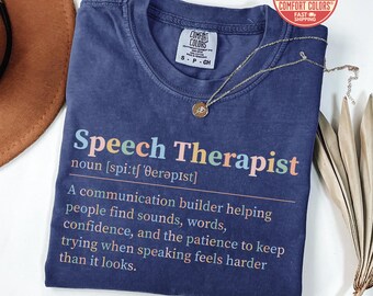 May include: Navy blue Comfort Colors t-shirt with the words "Speech Therapist" in colorful text. The definition of a speech therapist is printed below. A gold necklace with a pendant is draped on the shirt.