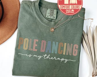 May include: A moss green t-shirt with the text "POLE DANCING is my therapy" in colorful letters. A gold necklace with a pendant rests on the shirt. The shirt has a Comfort Colors label.
