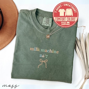 May include: An olive green t-shirt with the words "milk machine 24/7" in pastel colors. A gold necklace and a Comfort Colors logo are also visible. The shirt is folded on a white surface.