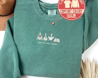 May include: Light green Comfort Colors sweatshirt with the text "just one more chapter." The sweatshirt features an embroidered design of figures reading books and a goose. A gold necklace and a brown hat are visible.