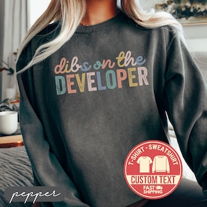 May include: Dark gray sweatshirt with the text "dibs on the DEVELOPER" in pastel colors. A red circular graphic with "T-SHIRT SWEATSHIRT CUSTOM TEXT FAST SHIPPING" is on the lower right. The word "pepper" is written in cursive on the lower left.