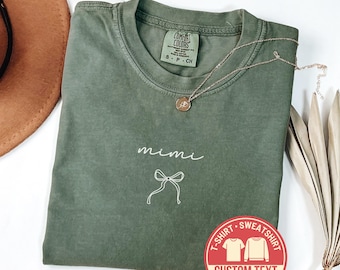 Puede incluir: Camiseta Comfort Colors verde musgo con la palabra "mimi" y un lazo. Un collar dorado con un colgante descansa sobre la camisa. Una pegatina roja con "Custom Text" es visible.