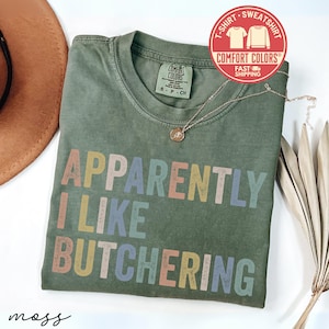 May include: An olive green t-shirt with the text "APPARENTLY I LIKE BUTCHERING" in colorful block letters. The shirt has a crew neck and short sleeves. A gold necklace with a pendant is draped on the shirt. A red and white sticker is in the upper right corner.