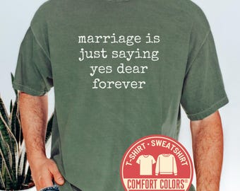 May include: Moss green t-shirt with the text "marriage is just saying yes dear forever" in white. The shirt has a Comfort Colors label and a red circular graphic with the words "T-shirt Sweatshirt".
