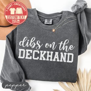 May include: A dark gray sweatshirt with the text "dibs on the DECKHAND" in white. The sweatshirt has a crew neck and long sleeves. A small gold necklace is visible. A red circle with text is in the upper left corner.