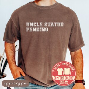 May include: Espresso-colored t-shirt with the text "UNCLE STATUS: PENDING" in a distressed white font. The shirt features a Comfort Colors label and a fast shipping badge. The model is wearing the shirt.