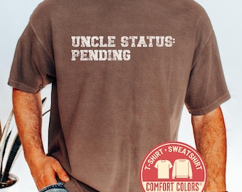 May include: Espresso-colored t-shirt with the text "UNCLE STATUS: PENDING" in a distressed white font. The shirt features a Comfort Colors label and a fast shipping badge. The model is wearing the shirt.