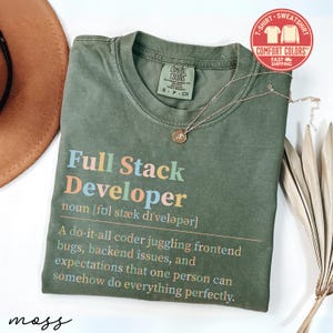 May include: Olive green t-shirt with the text "Full Stack Developer" in colorful letters. The definition of a full stack developer is printed below the title. A gold necklace and a brown hat are also in the image.