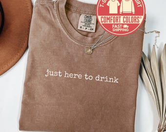May include: Espresso-colored t-shirt with the phrase "just here to drink" in white lettering. The shirt has a crew neck and a Comfort Colors tag. A silver necklace with a pendant is draped across the shirt. A red and white circular graphic indicates the shirt is fast shipping.