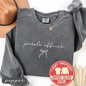 May include: A dark gray Comfort Colors sweatshirt with the words "parole officer" and a bow graphic in white script. A gold necklace and a brown hat are visible. A red and white Comfort Colors logo is on the right.