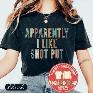 May include: Black t-shirt with the text "APPARENTLY I LIKE SHOT PUT" in colorful block letters. The shirt has a Comfort Colors logo and the word "black" in a cursive font.