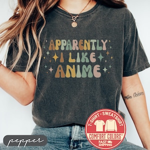 May include: Dark gray t-shirt with the text "APPARENTLY I LIKE ANIME" in retro-style lettering. The design features various colors, including brown, yellow, green, and pink. The shirt has a Comfort Colors tag.