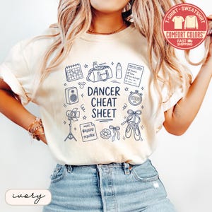 May include: An ivory t-shirt featuring a dark blue graphic print with the text "DANCER CHEAT SHEET" surrounded by illustrations of a calendar, gym bag, water bottle, practice notes, speaker, stopwatch, stage light, music sheet, scrunchie, and ballet shoes.