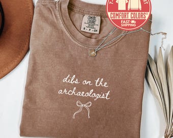 May include: Espresso-colored t-shirt with the phrase "dibs on the archaeologist" in white script. A silver necklace with a pendant rests on the shirt. The shirt has a Comfort Colors label.