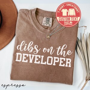 May include: Espresso-colored t-shirt with the phrase "dibs on the DEVELOPER" in white lettering. The shirt has a crew neck and short sleeves. A necklace with a small pendant is draped on the shirt. A circular graphic reads "CUSTOM TEXT" and "FAST SHIPPING".