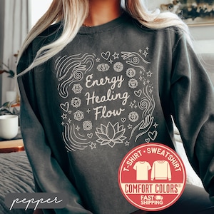 May include: A dark gray sweatshirt with the words "Energy Healing Flow" in white script, surrounded by line art of hearts, stars, hands, and a lotus flower. The sweatshirt has a Comfort Colors label.