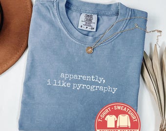 May include: A blue jean Comfort Colors t-shirt with the phrase "apparently, i like pyrography" in white lettering. A gold necklace with a pendant rests on the shirt. A brown hat is visible in the background.