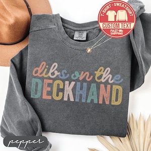 May include: A dark gray sweatshirt with the phrase "dibs on the DECKHAND" in colorful letters. The sweatshirt has a crew neck and long sleeves. A red and white sticker with the words "CUSTOM TEXT" is visible.
