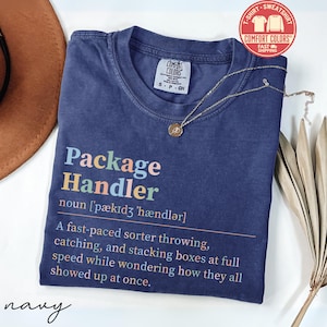 May include: Navy blue t-shirt with the text "Package Handler" in colorful letters. The definition of "Package Handler" is printed below the title. A gold necklace with a pendant is draped on the shirt.