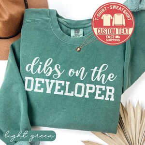 Puede incluir: Una sudadera verde claro con la frase "dibs on the DEVELOPER" en letras blancas. La sudadera tiene cuello redondo y mangas largas. Se ve un collar dorado con un colgante. Un gráfico circular rojo con texto está en la esquina superior derecha.